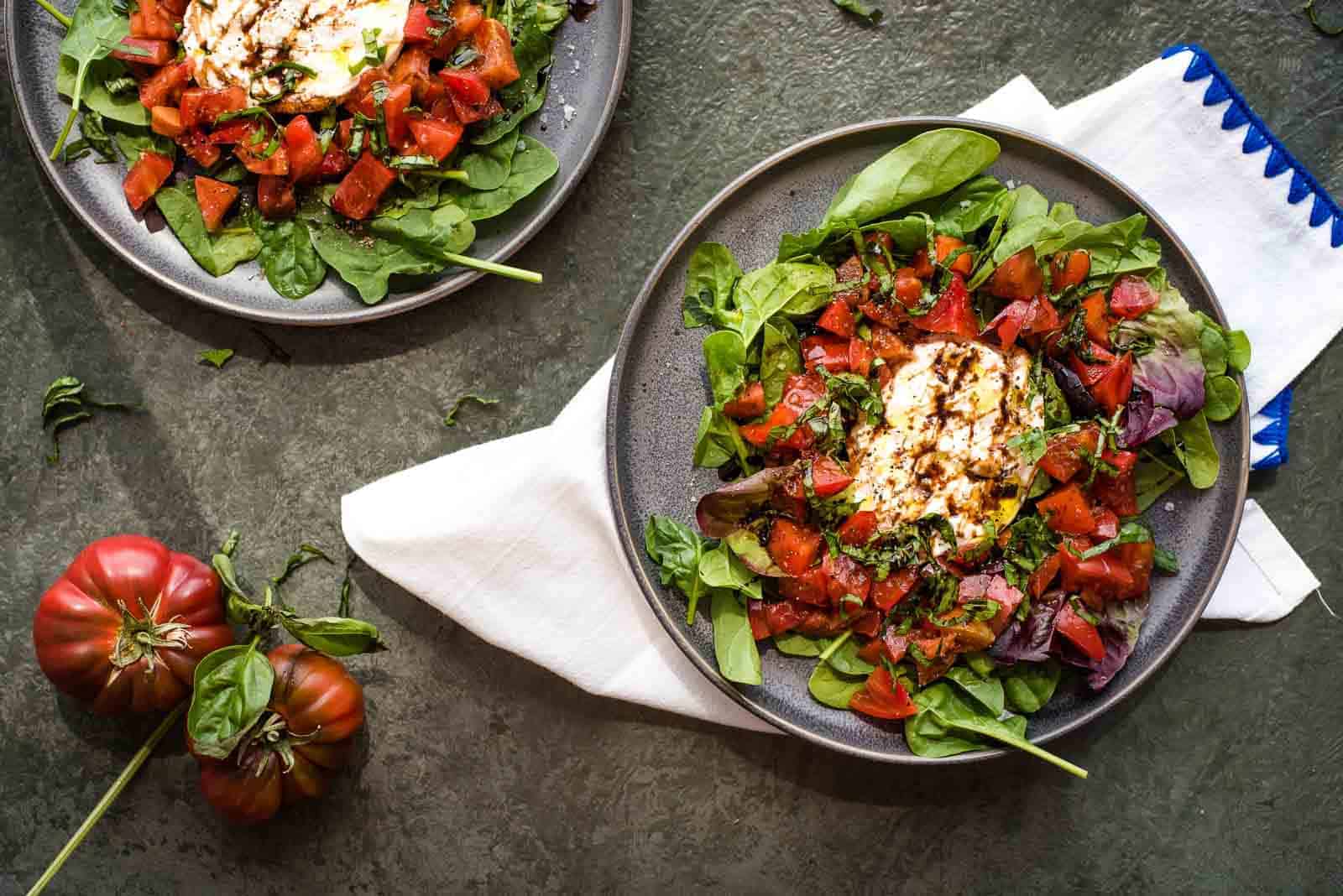 Two plates of salad with fresh greens, diced tomatoes, burrata cheese, and balsamic glaze, placed on a dark surface next to two tomatoes and a napkin.
