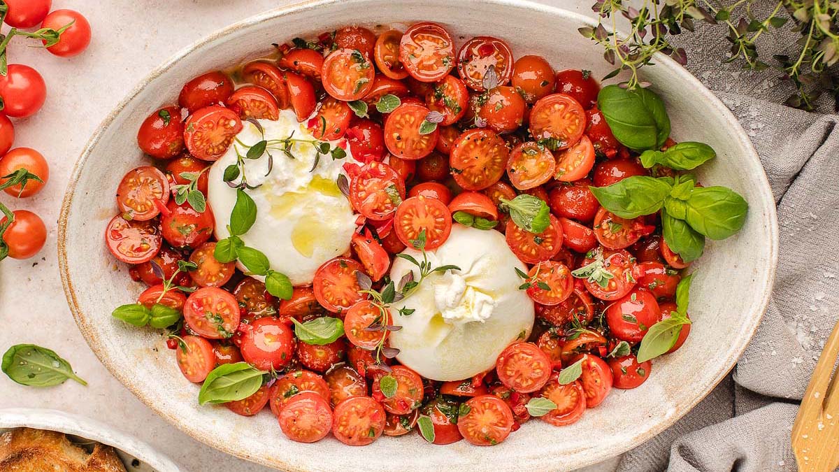 Oval platter with cherry tomato halves, two balls of burrata cheese, fresh basil leaves, and herbs, drizzled with olive oil.