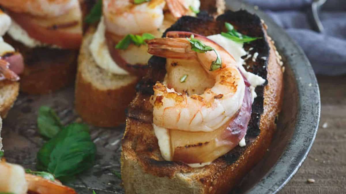 Toasted bread slices topped with shrimp, bacon, and a dollop of creamy spread, garnished with fresh herbs, on a metal tray.