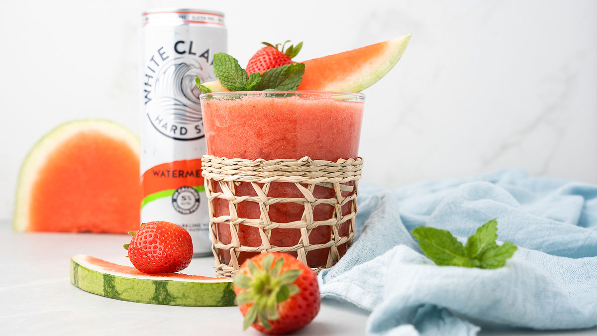 A glass of watermelon slush garnished with a watermelon slice, strawberry, and mint sits in front of a White Claw can, with fruit and a blue cloth nearby.