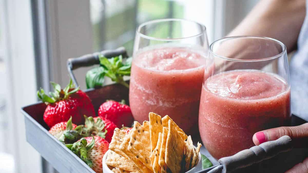 A tray holding two glasses of pink frozen drink, several fresh strawberries, green mint, and a stack of crackers in a small bowl.