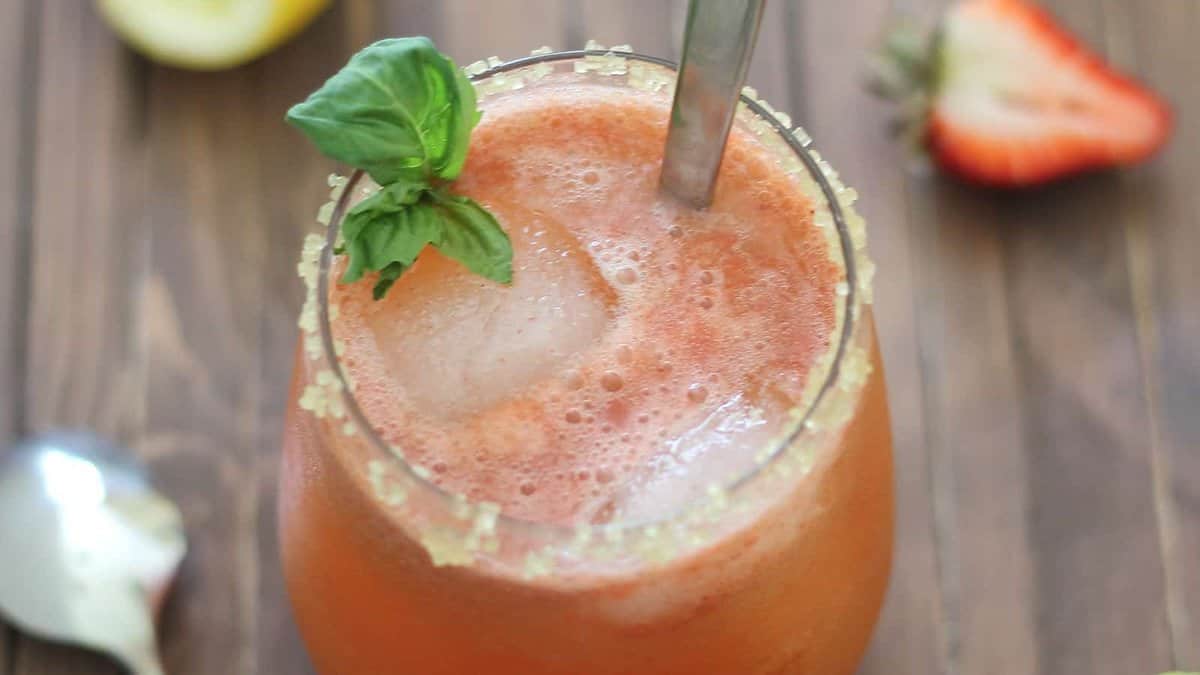 A glass of iced strawberry drink with a sugar rim and basil garnish, a metal straw, and a strawberry and lemon slice in the background on a wooden surface.