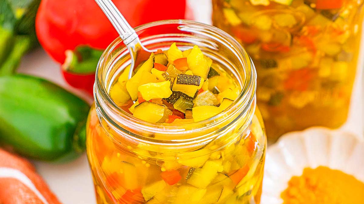 A glass jar filled with chopped pickled vegetables, including zucchini and peppers, with a spoon inside. Fresh vegetables and a jar are visible in the background.