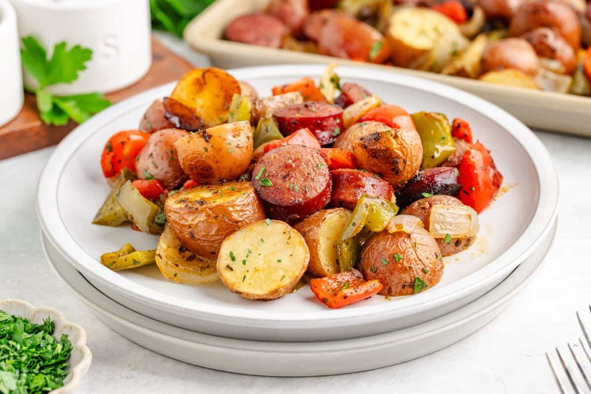 A plate of roasted potatoes, sliced sausage, bell peppers, and onions, garnished with herbs, sits on a white table next to a dish of chopped parsley.