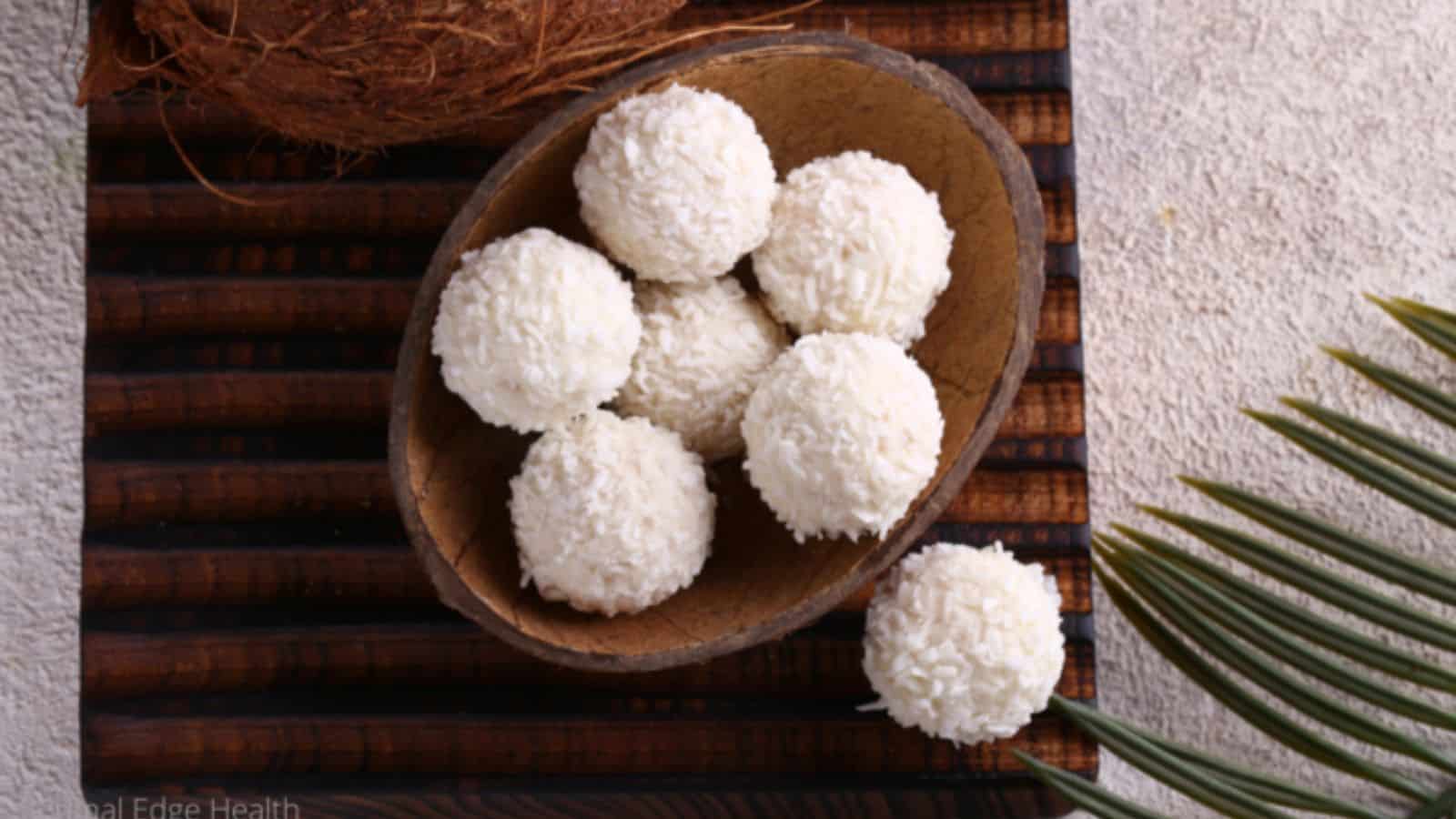 A wooden bowl filled with round, white coconut-covered sweets sits on a striped wooden mat, with a coconut shell and palm leaf nearby.
