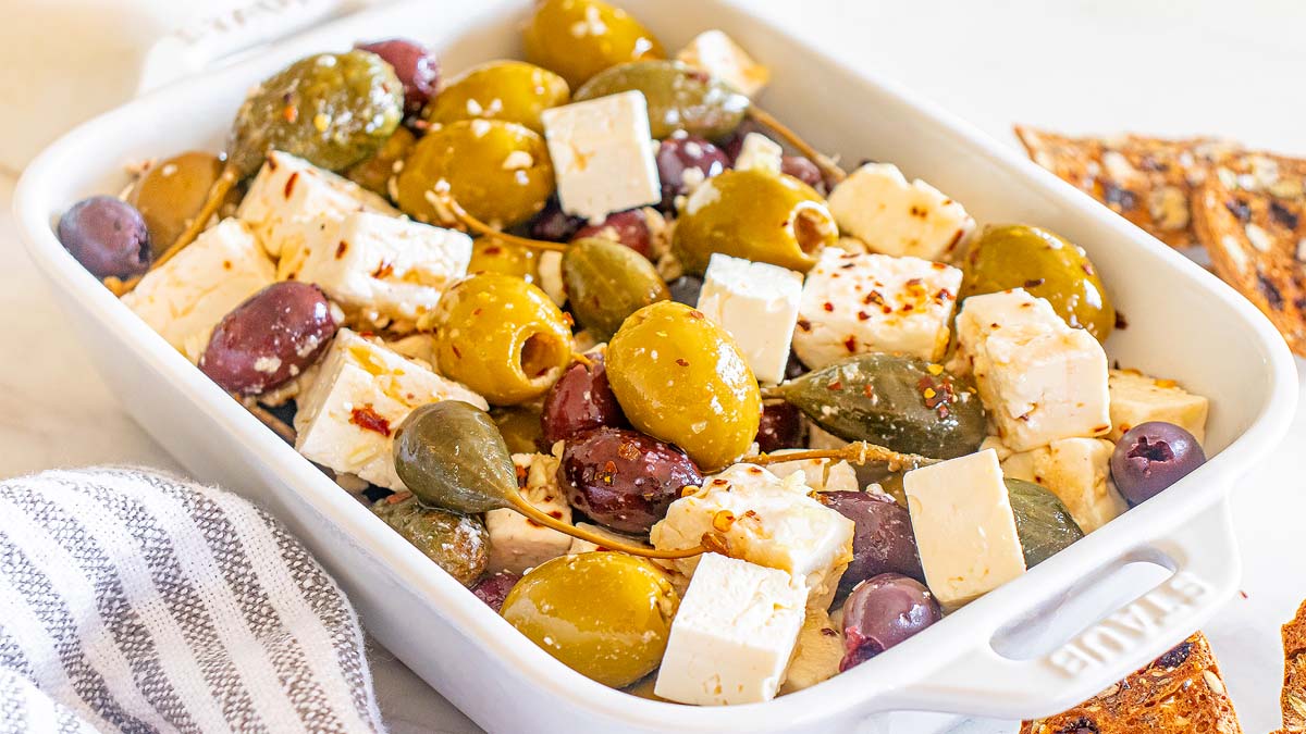 A white ceramic dish filled with mixed olives, caper berries, and cubes of feta cheese, seasoned with herbs and chili flakes.