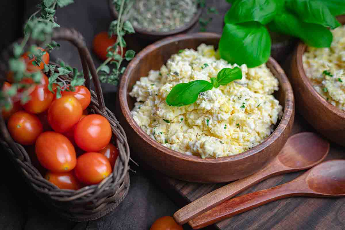 A wooden bowl filled with scrambled eggs garnished with fresh basil sits beside a basket of grape tomatoes and two wooden spoons.