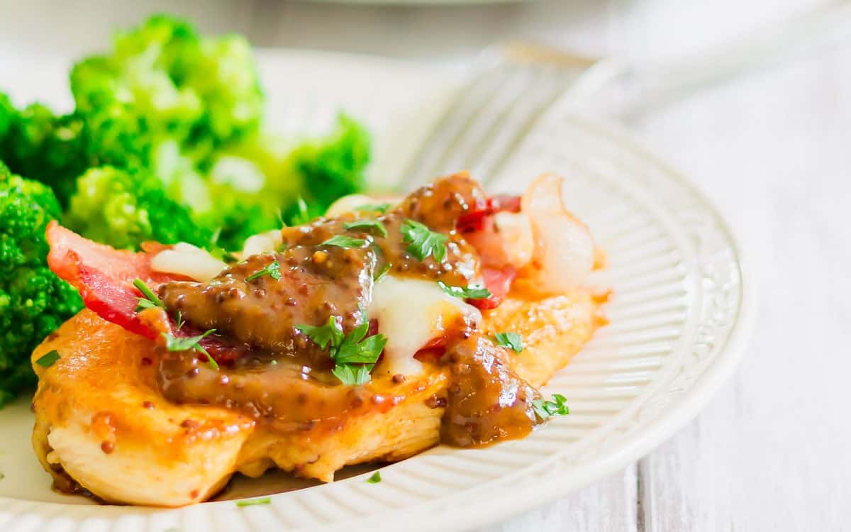 Grilled chicken breast topped with melted cheese, bacon, sauce, and herbs, served with a side of steamed broccoli on a white plate.