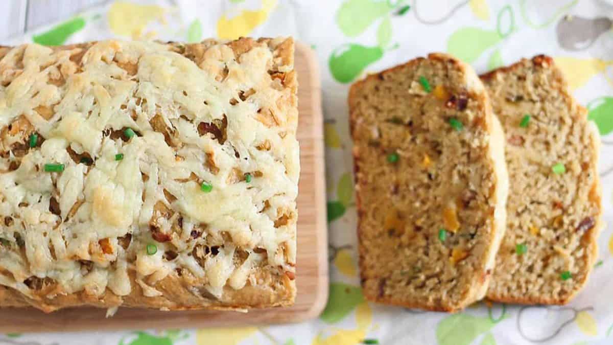 A loaf of savory bread topped with melted cheese and chopped chives, with two sliced pieces next to it on a patterned surface.