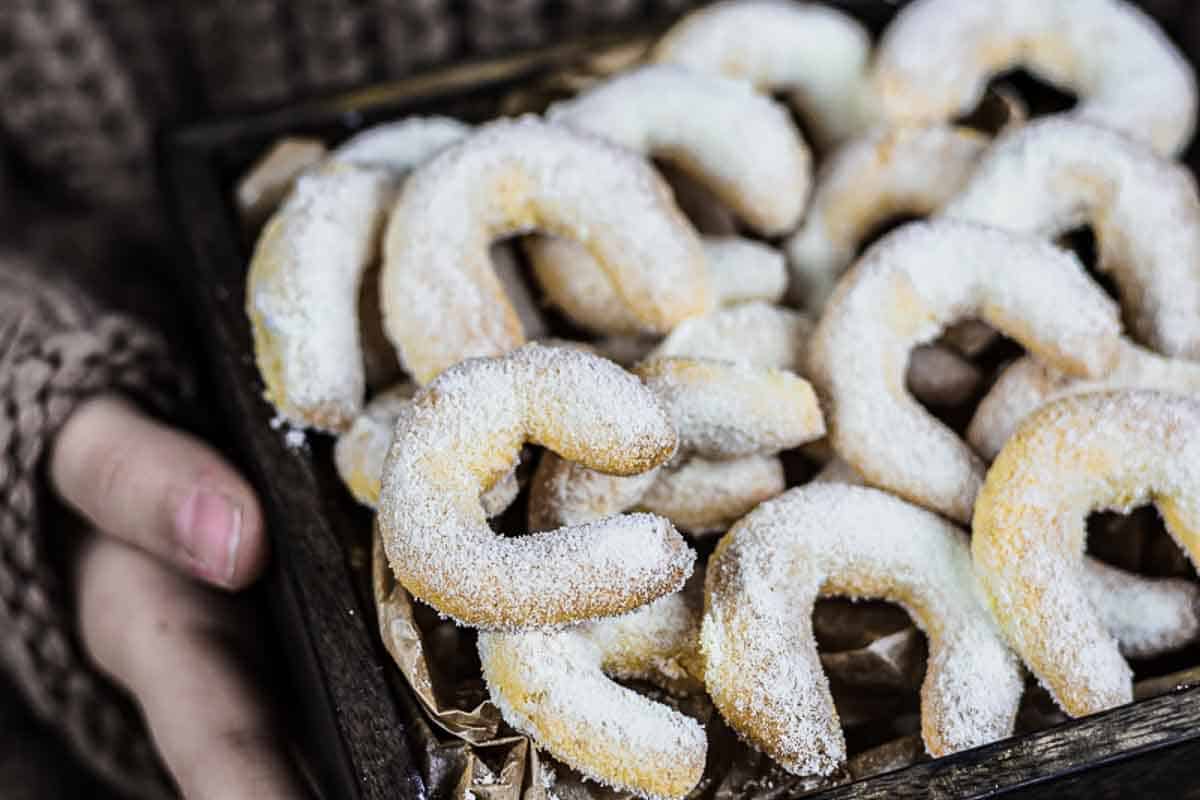 A person holding a tray of crescent-shaped cookies dusted with powdered sugar.