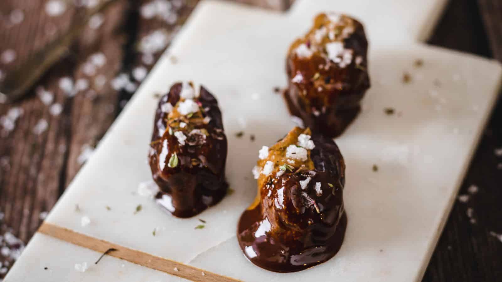 Three dates coated in chocolate and sprinkled with sea salt are arranged on a white marble board.