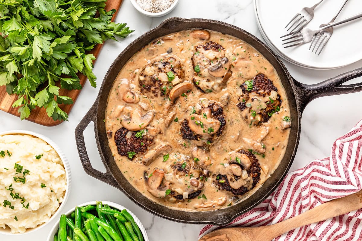 A skillet with Salisbury steak patties in mushroom gravy, surrounded by bowls of mashed potatoes, green beans, parsley, and plates with utensils.