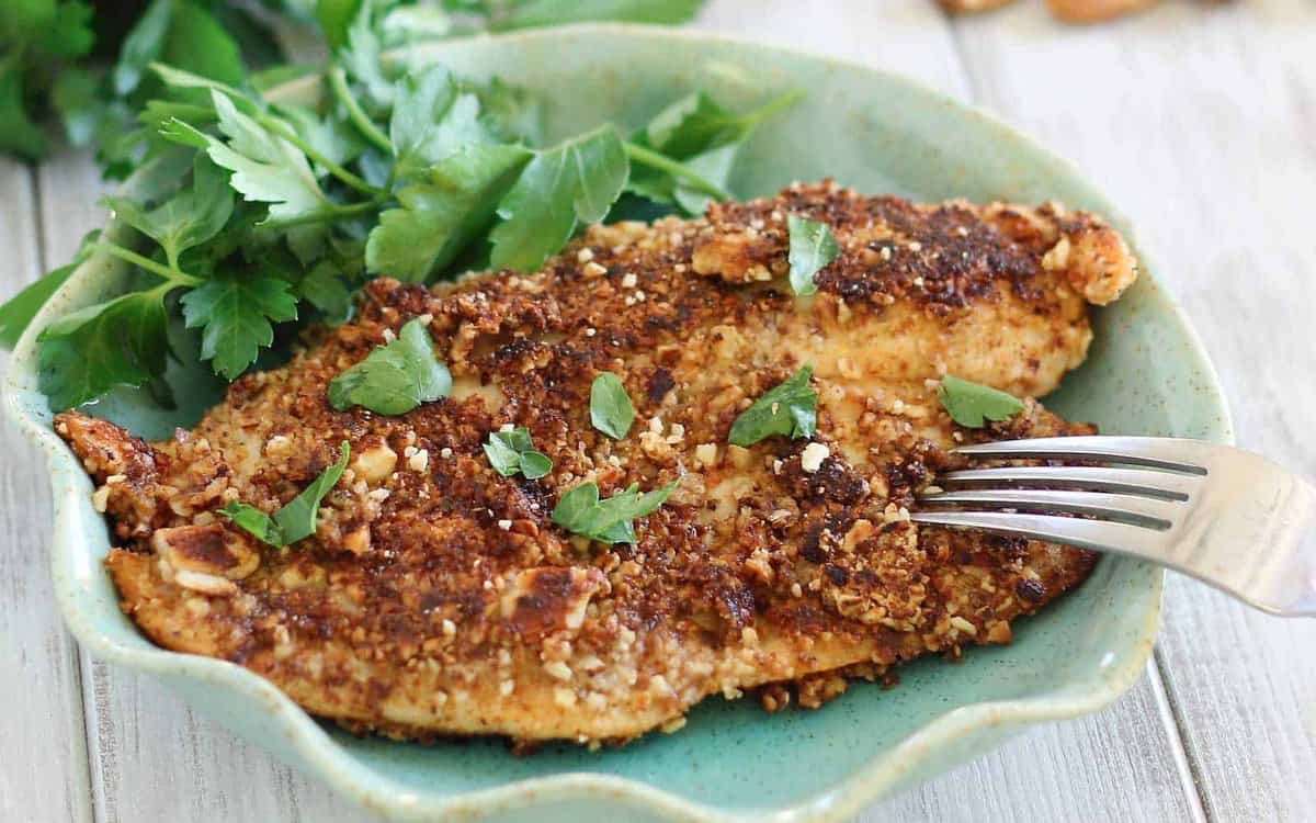 A cooked, breaded fish fillet topped with fresh herbs sits on a green plate next to a sprig of parsley; a fork is placed on the fish.