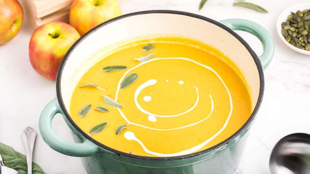 A pot of creamy orange soup garnished with a swirl of cream and sage leaves sits on a white surface with apples and a bowl of seeds nearby.