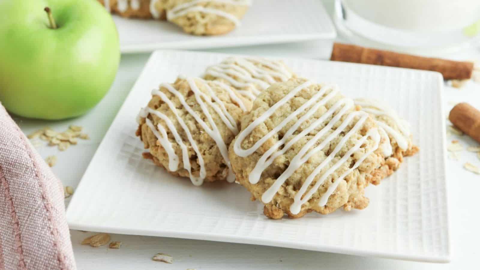 Three apple oatmeal cookies with white icing drizzle are arranged on a square white plate, with a green apple and cinnamon sticks in the background.