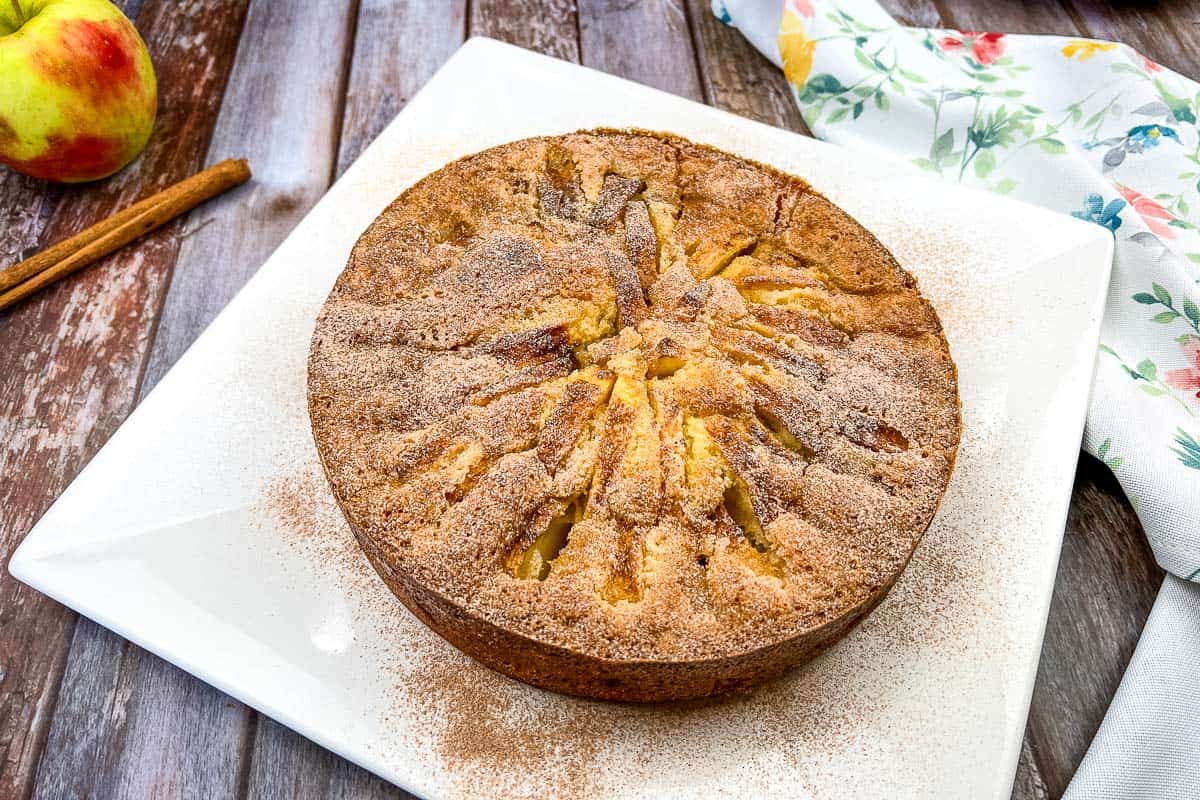 A round apple cake topped with cinnamon sugar sits on a white square plate, with a cinnamon stick, apple, and floral napkin nearby on a wooden table.