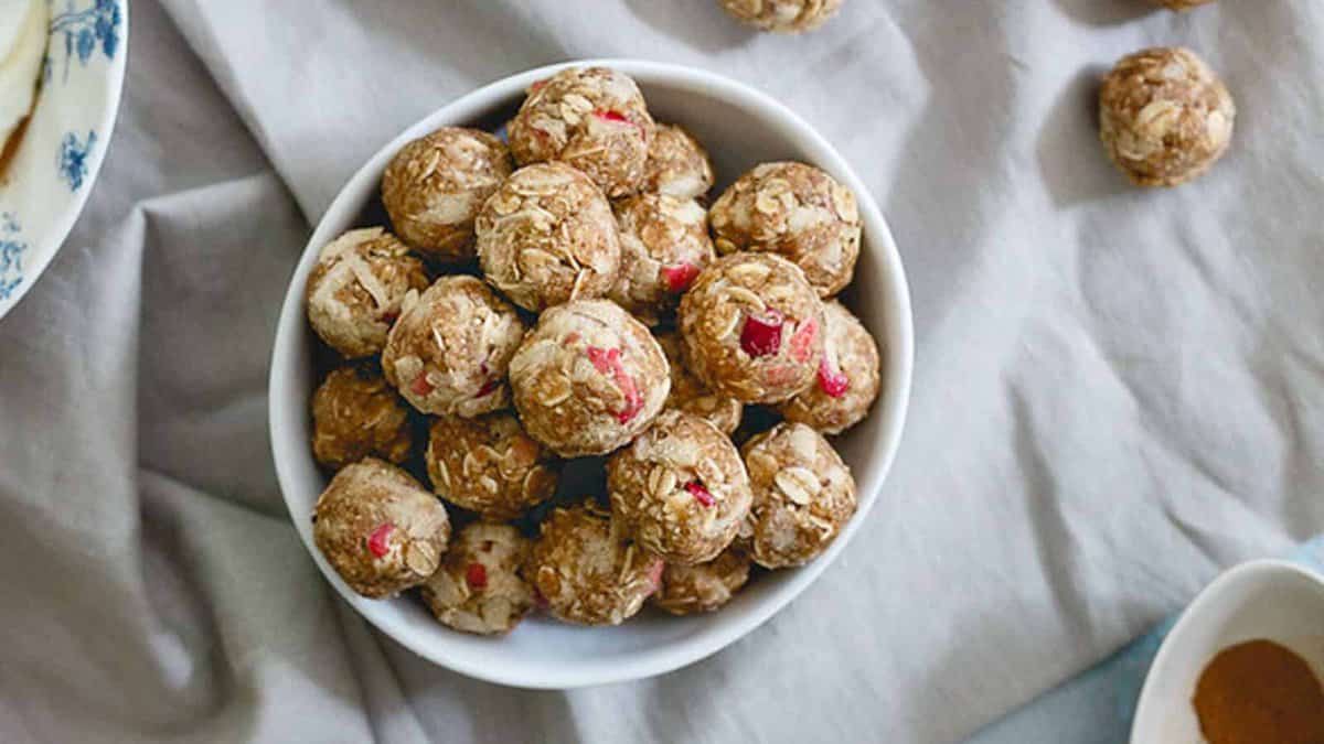 A white bowl filled with round oat and fruit energy bites sits on a light-colored cloth, with a few bites scattered nearby.