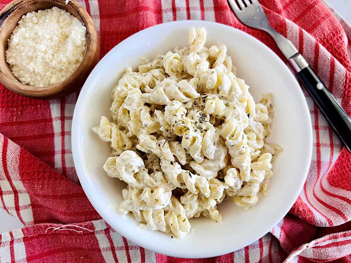 A white bowl of creamy rotini pasta sits on a red and white checkered cloth, with a fork and a small wooden bowl of grated cheese nearby.
