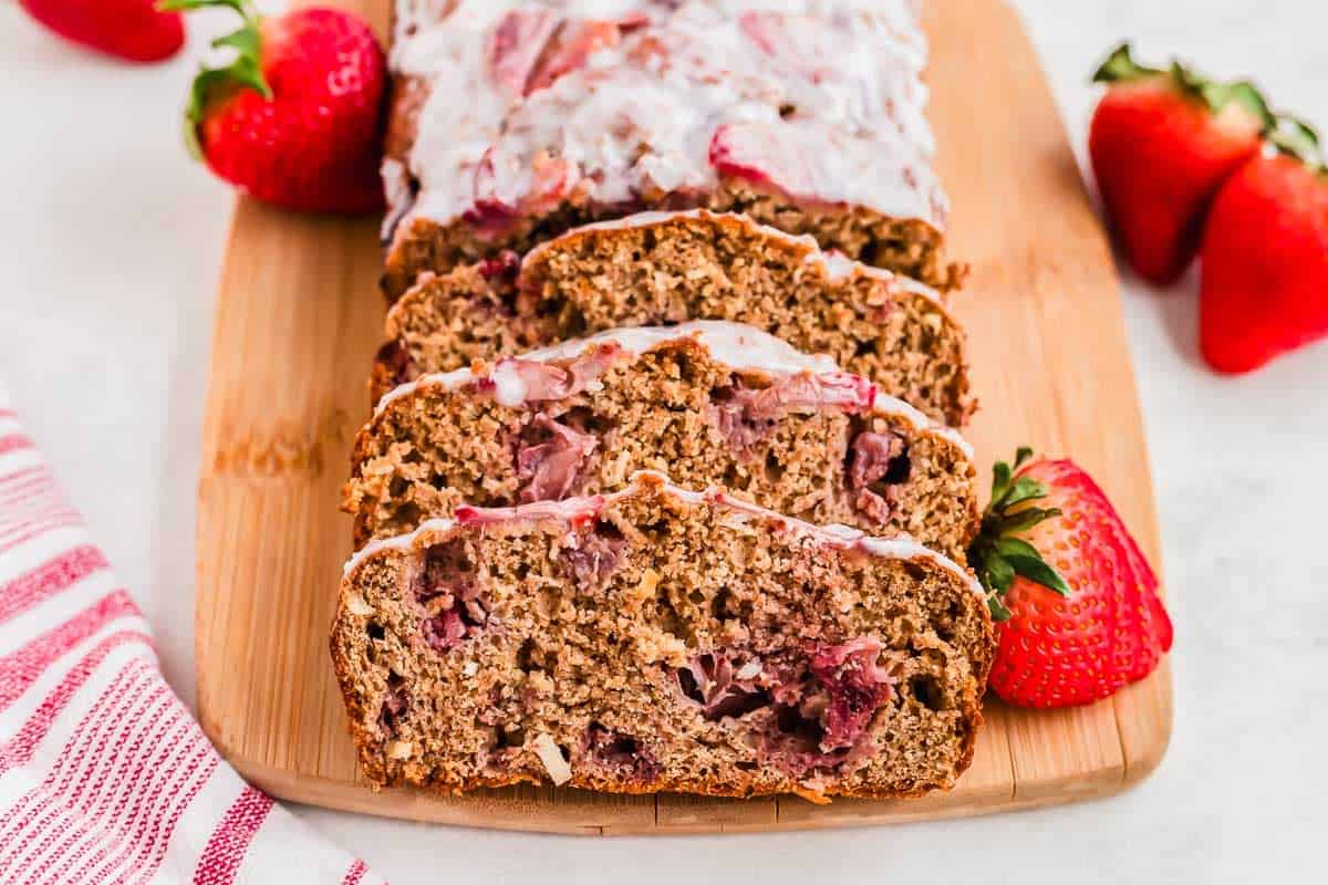 Sliced strawberry bread with icing on a wooden cutting board, surrounded by fresh strawberries.