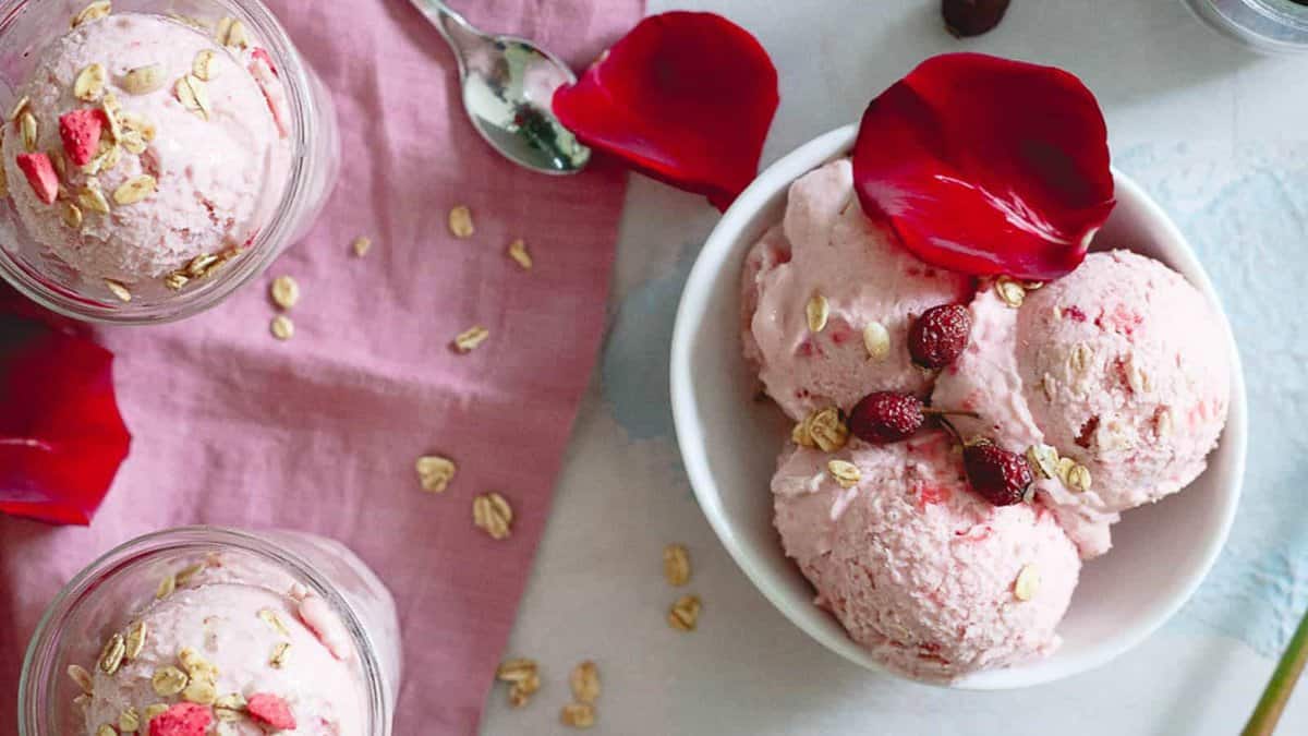 A bowl of pink ice cream garnished with oats, dried berries, and rose petals sits next to a spoon and two jars of similar ice cream on a light surface with a pink cloth.