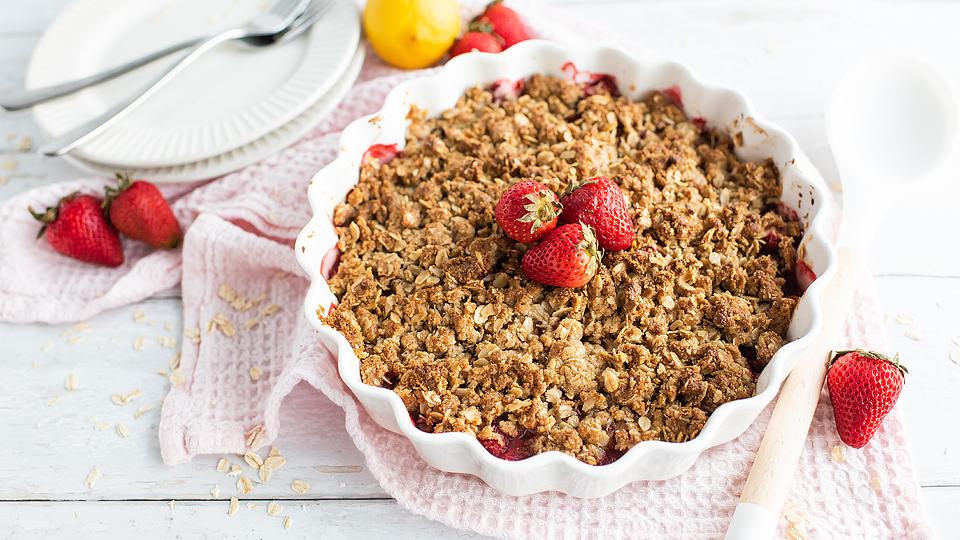 A baked strawberry crumble topped with fresh strawberries in a white dish, surrounded by whole strawberries, plates, and utensils on a light pink cloth.