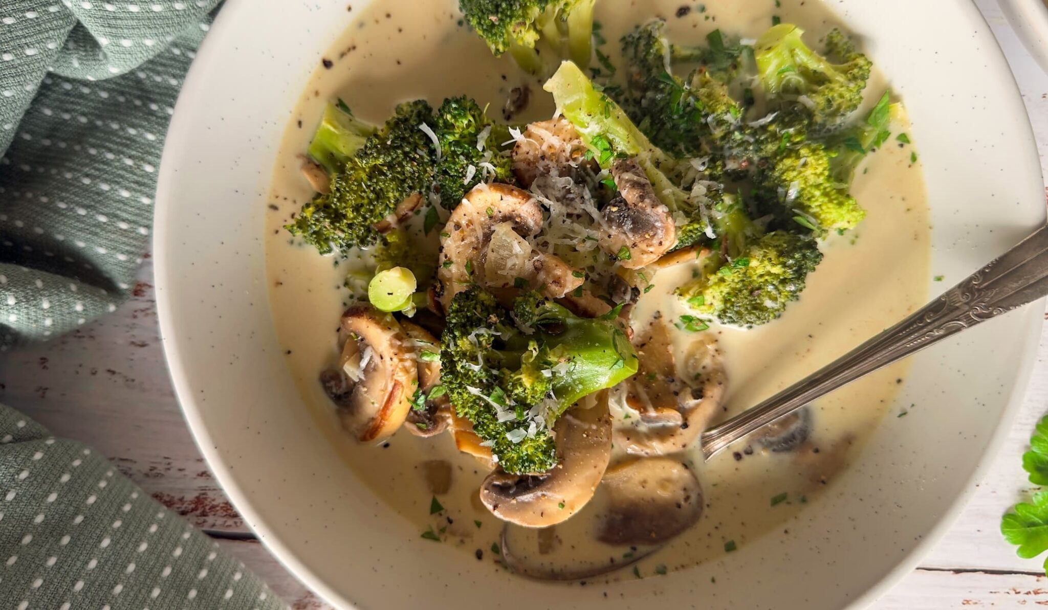 A bowl of creamy soup with broccoli, sliced mushrooms, chopped green onions, and herbs, with a spoon resting in the bowl.