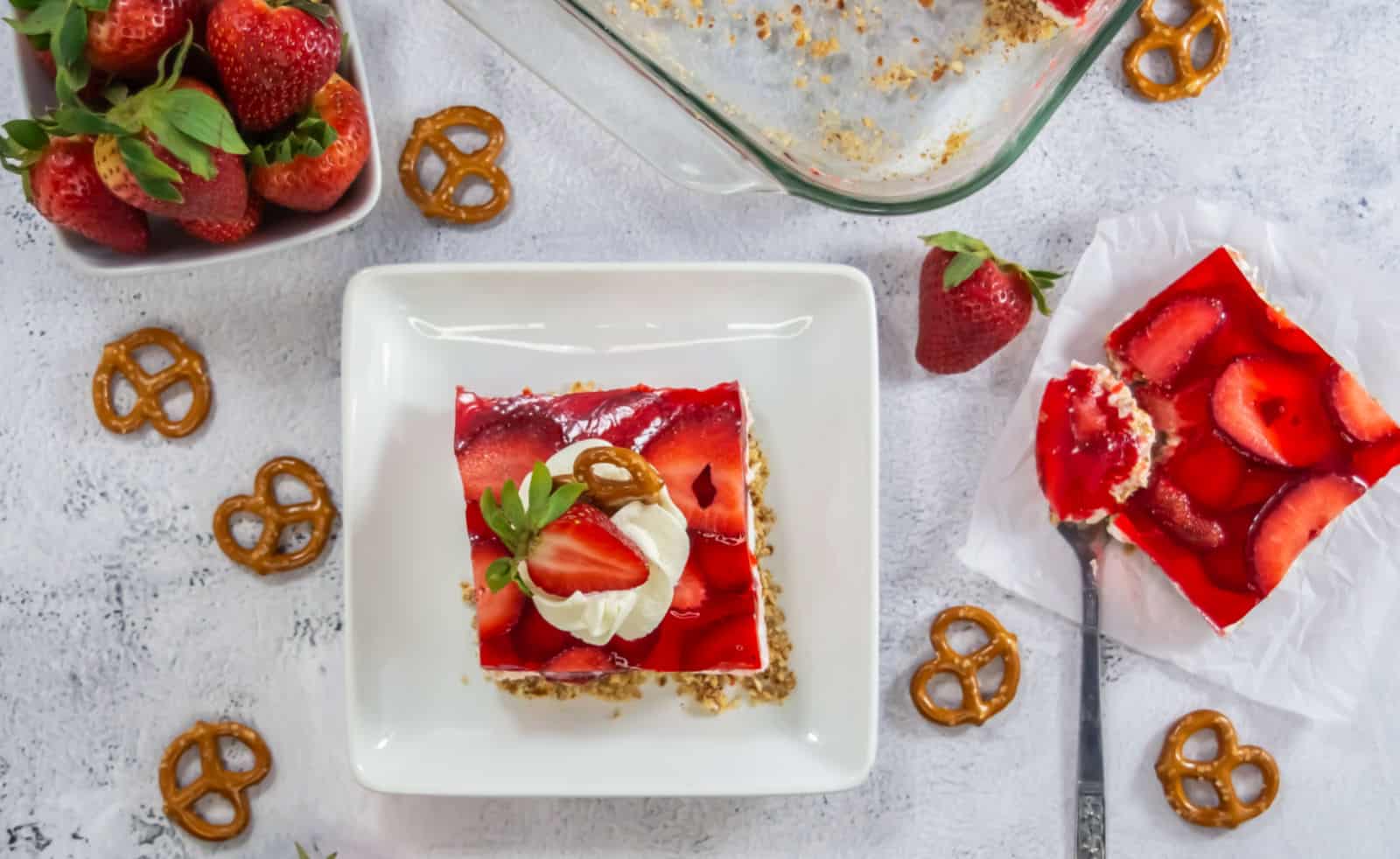 A square slice of strawberry pretzel dessert on a white plate, topped with whipped cream, a pretzel, and fresh strawberries, with extra dessert, strawberries, and pretzels nearby.
