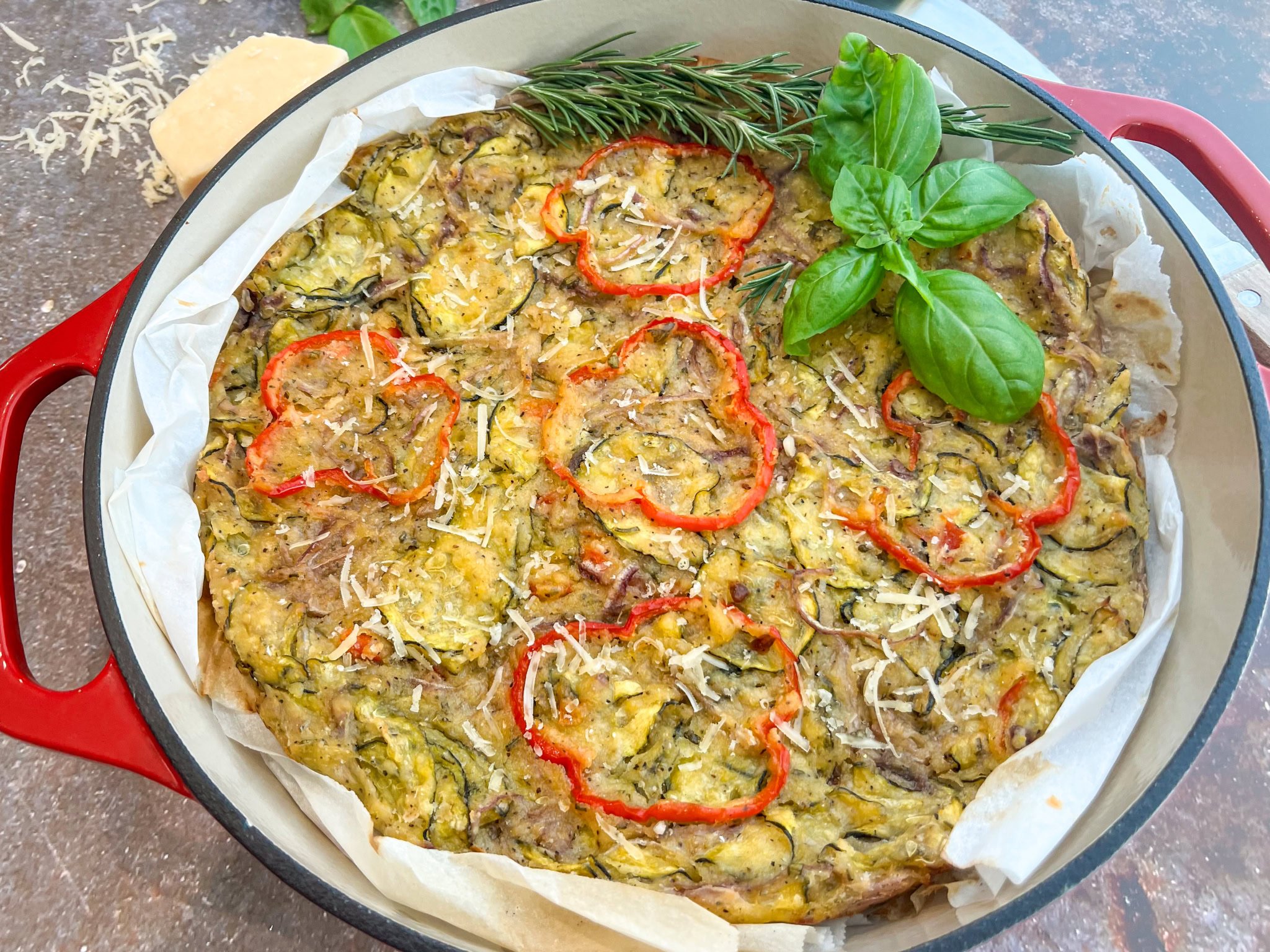 Baked vegetable frittata topped with red bell pepper slices, grated cheese, rosemary, and basil, served in a round red baking dish lined with parchment paper.