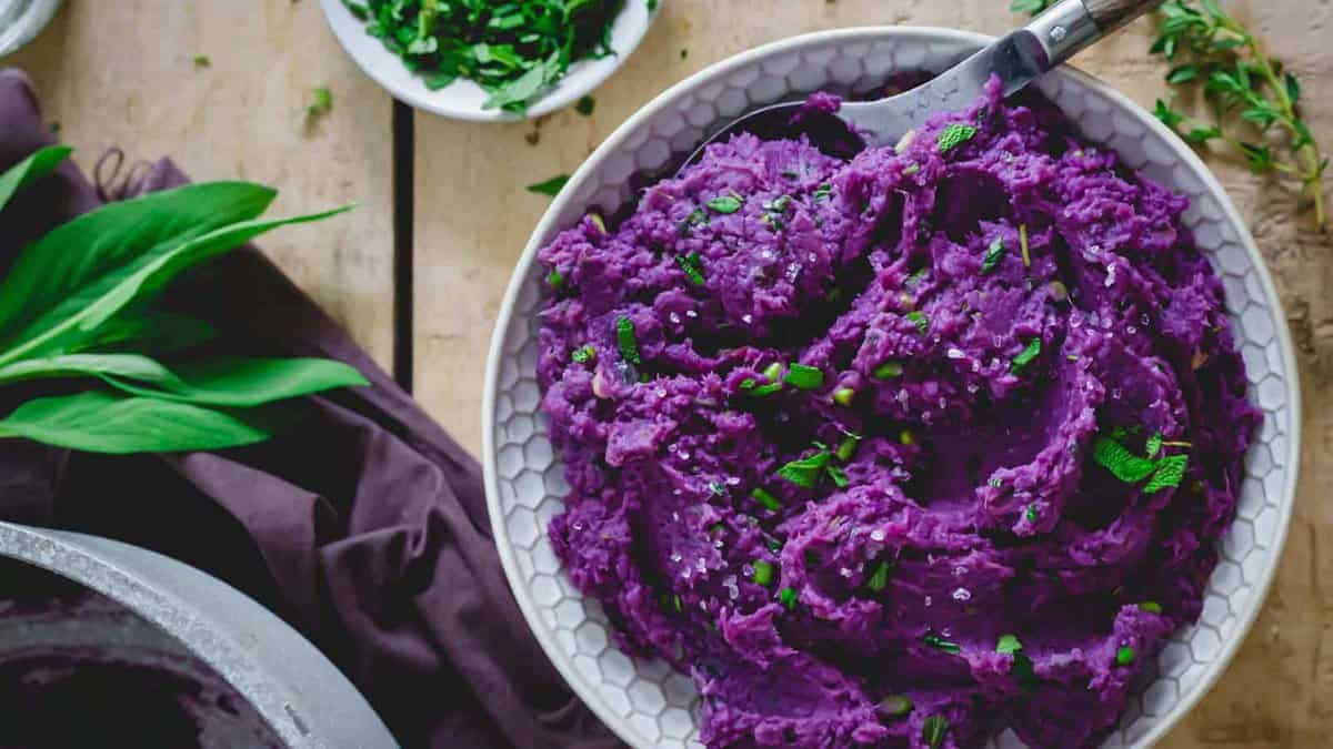 Bowl of mashed purple sweet potatoes garnished with chopped herbs, with a spoon and fresh herbs on the side.