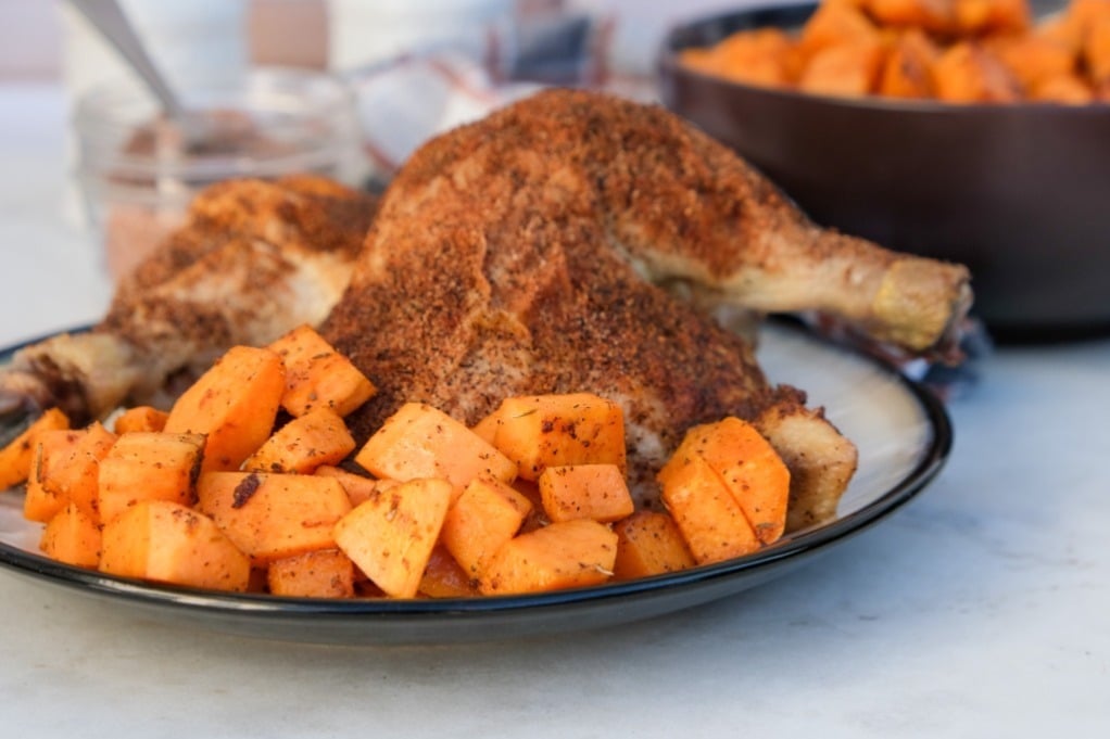 A roasted whole chicken with a seasoning crust is served on a plate with a side of roasted sweet potato cubes.