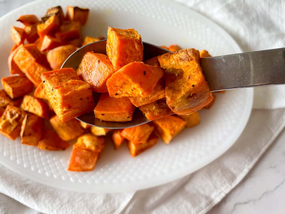 A spoon holds several roasted sweet potato cubes above a white plate with more roasted sweet potato cubes on it.
