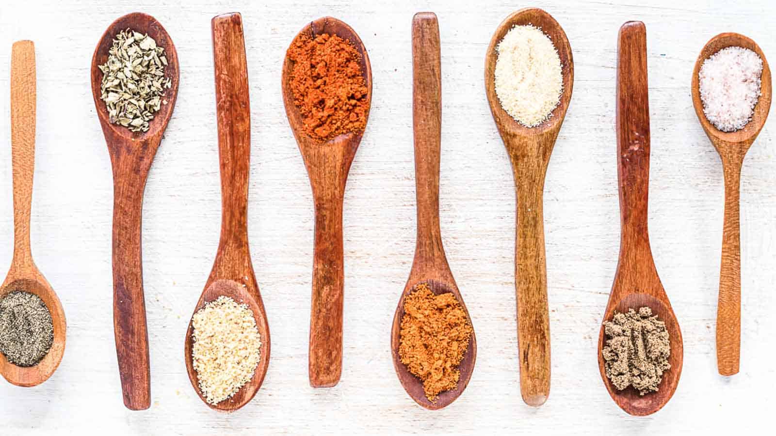 Eight wooden spoons on a white surface, each holding a different spice or seasoning, arranged in a row.