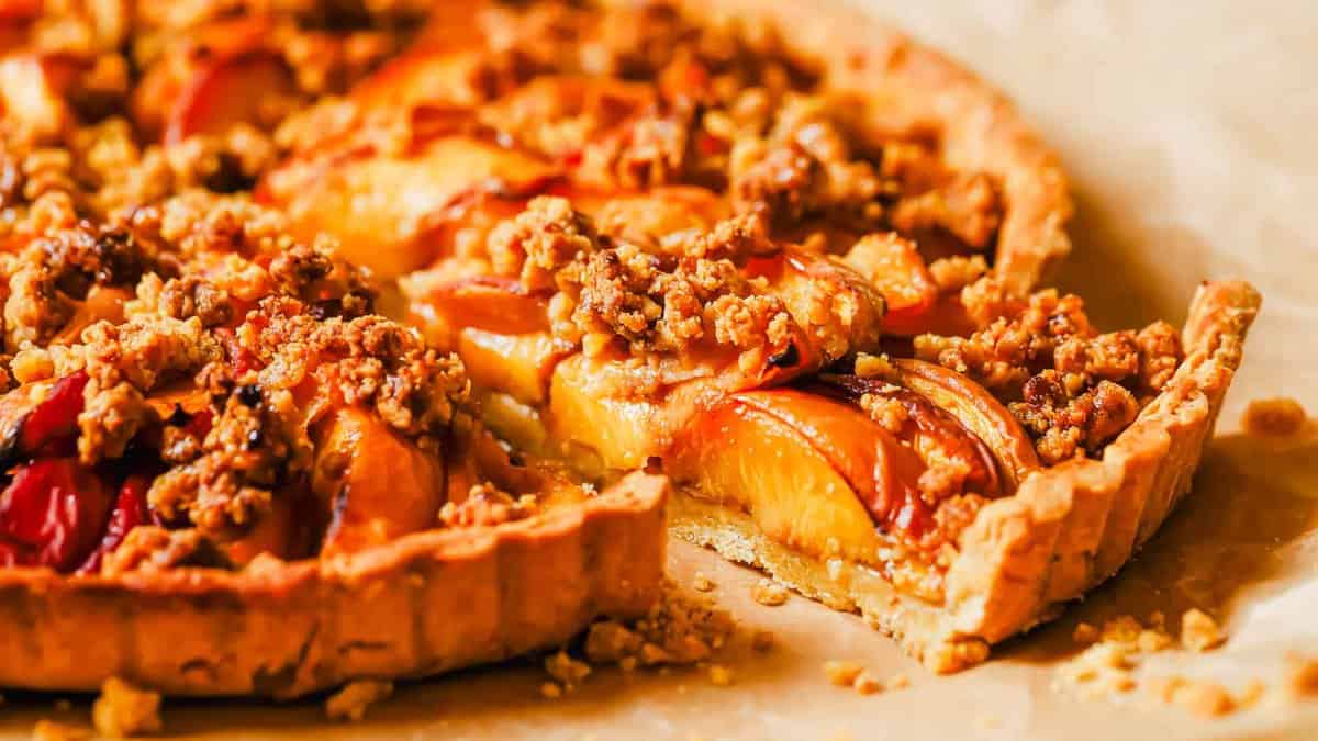 A close-up of a peach crumble tart with a slice partially removed, showing the baked fruit filling and crumbly topping.