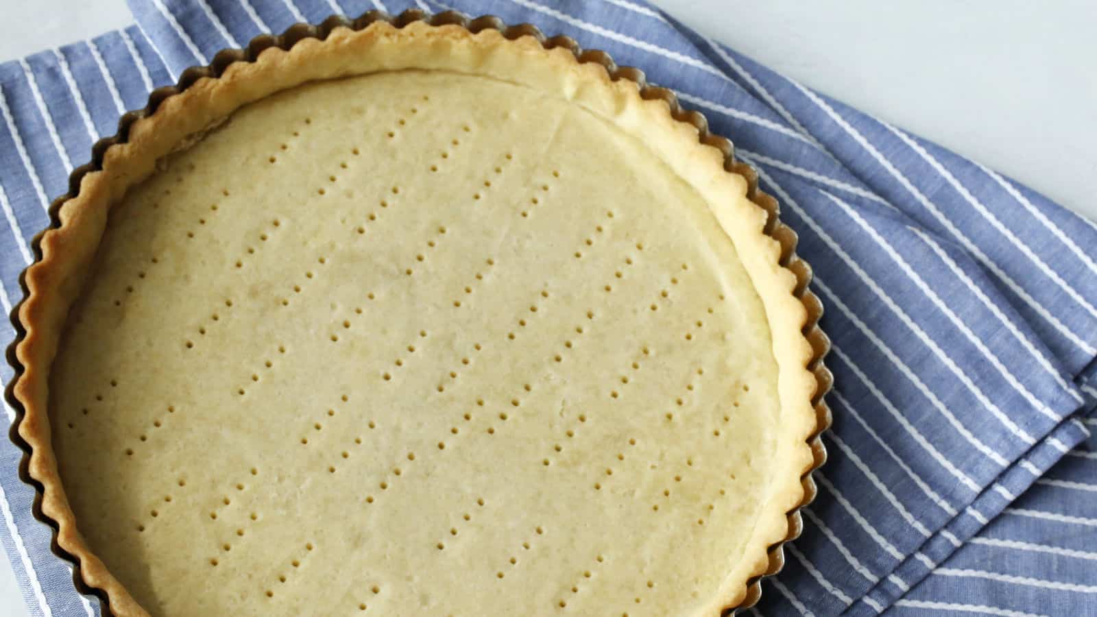 Unbaked tart crust with fork marks sits in a fluted tart pan on a blue and white striped kitchen towel.