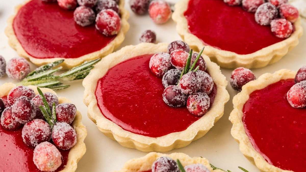 Mini tarts with smooth red filling, topped with sugared cranberries and sprigs of rosemary, arranged on a light surface.