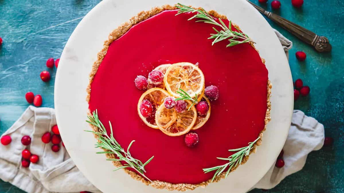 A cranberry tart with a glossy red surface, garnished with dried orange slices, cranberries, and sprigs of rosemary, sits on a white plate.