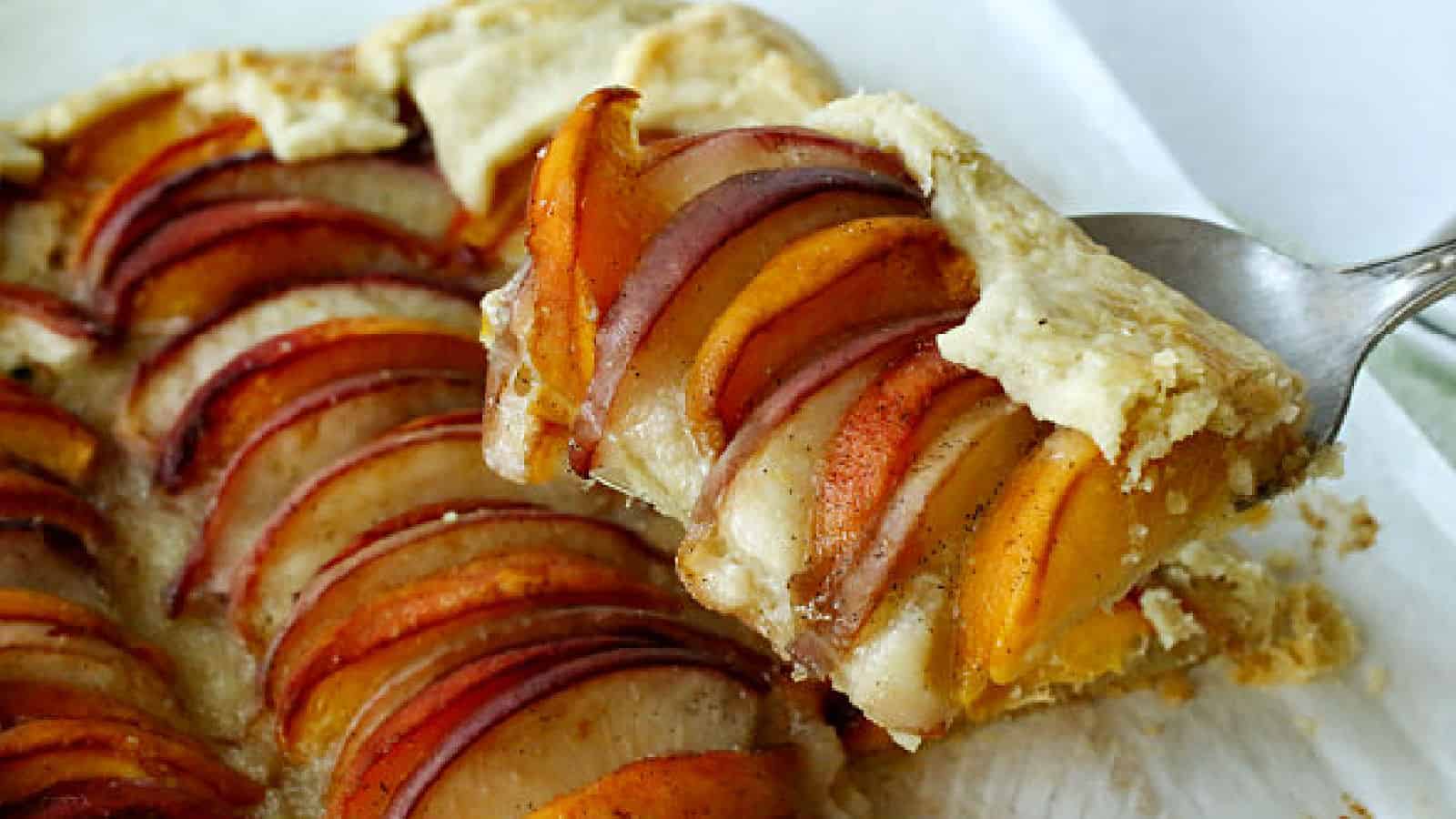 A slice of rustic fruit tart with thinly sliced plums and peaches on a flaky pastry crust is being lifted from a pan with a spatula.