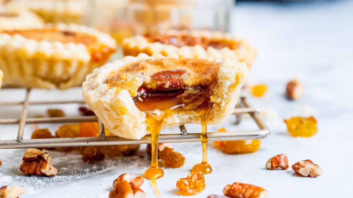 A butter tart with a bite taken out, showing gooey caramel filling and pecans, sits on a wire rack with scattered raisins and pecans around it.