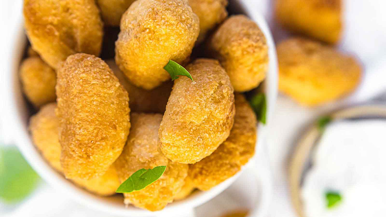 A bowl filled with golden-brown, crispy breaded snacks, garnished with small green leaves, sits next to a bowl of white dipping sauce.