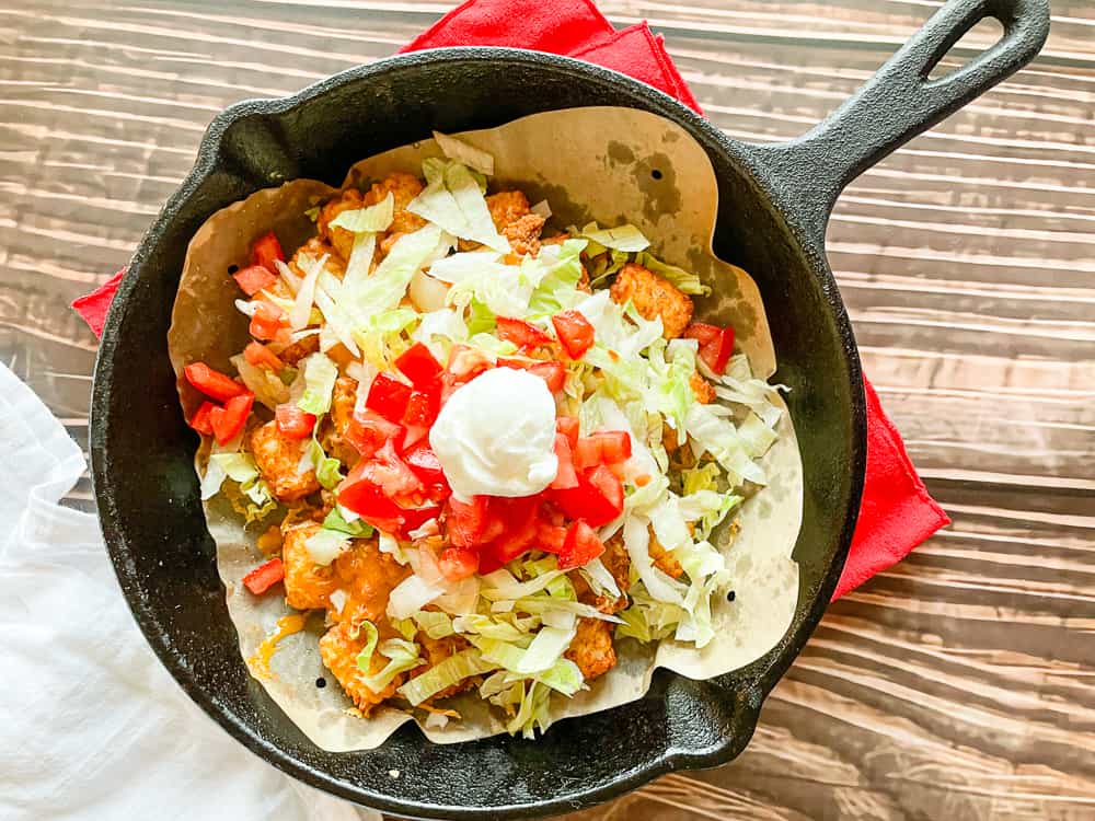 A cast iron skillet holds layered nachos topped with shredded lettuce, diced tomatoes, and a dollop of sour cream, resting on a red napkin.