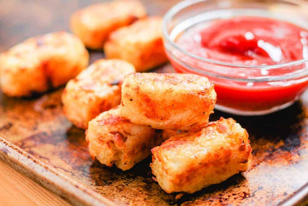 Several golden-brown tater tots are arranged on a tray next to a small glass bowl filled with ketchup.