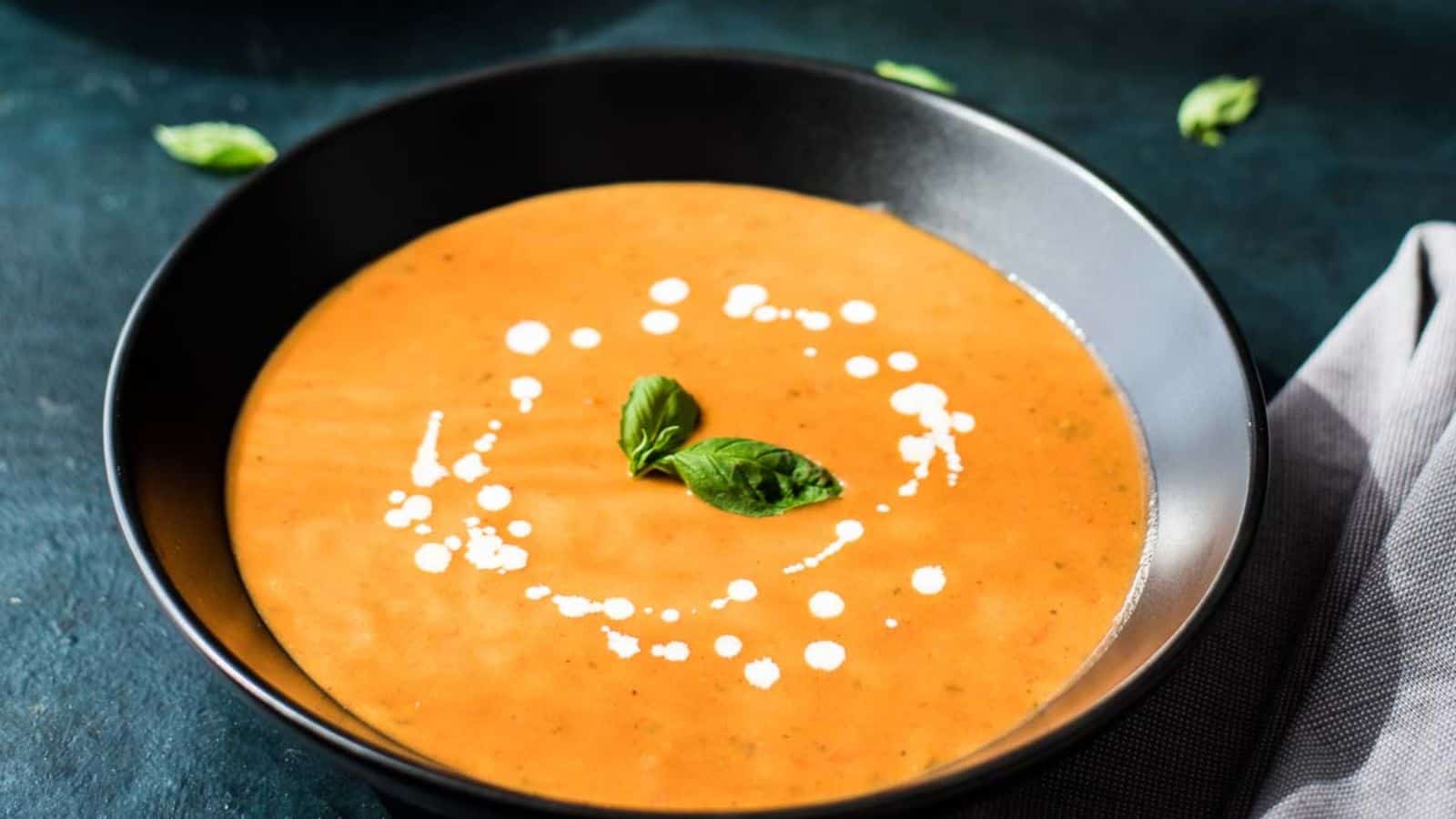 A bowl of creamy tomato soup garnished with a swirl of cream and fresh basil leaves, placed on a dark surface.