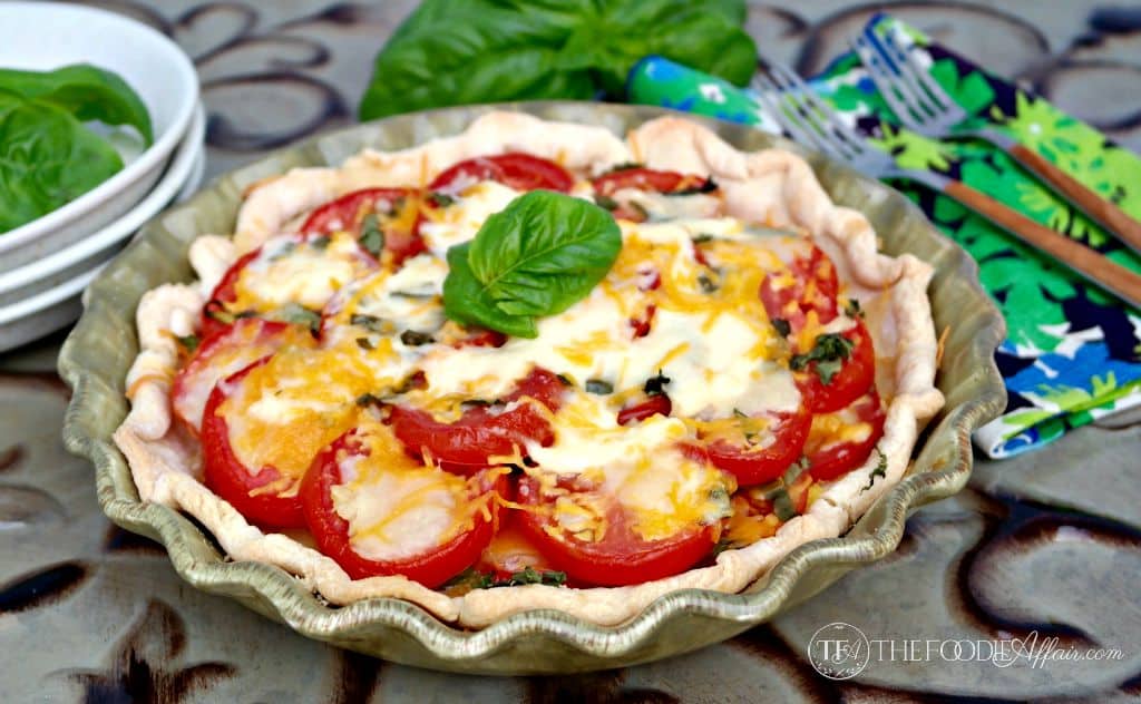 A baked tomato pie with melted cheese and basil, in a fluted pie dish, garnished with a fresh basil leaf. Plates and utensils are in the background.