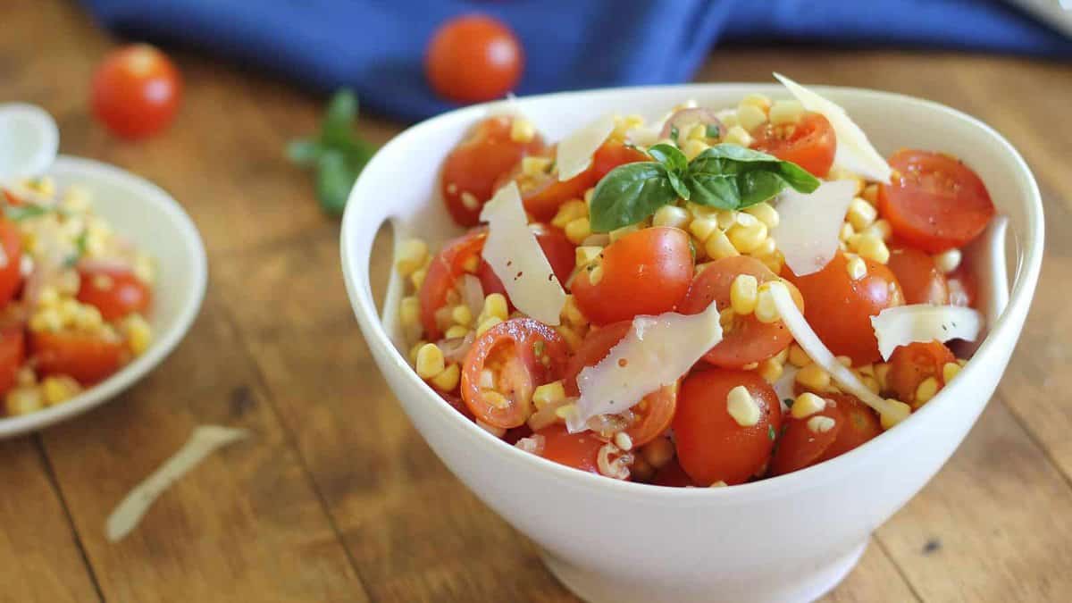A white bowl filled with a salad of cherry tomatoes, corn, shaved cheese, and basil sits on a wooden table.