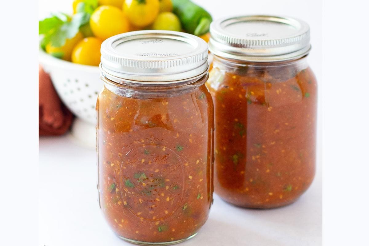 Two glass jars filled with homemade salsa sit on a white surface, with a bowl of yellow tomatoes and herbs in the background.