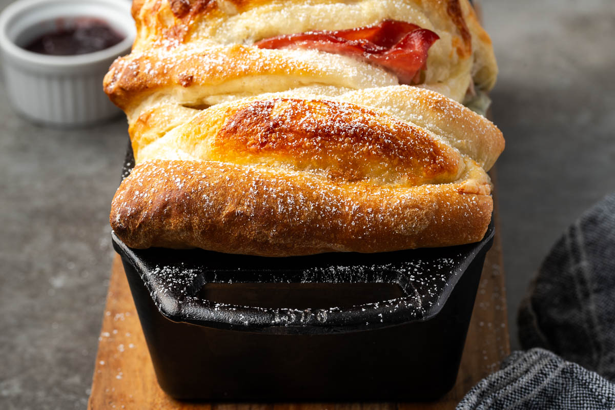 A loaf of braided bread baked in a black pan, lightly dusted with powdered sugar, sits on a wooden board with a small dish of jam in the background.