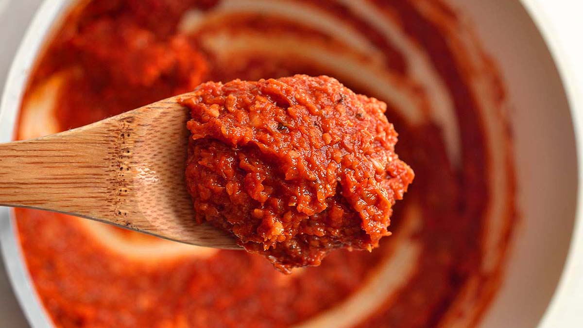 A wooden spoon holds a thick scoop of red tomato paste above a white bowl with more paste inside.