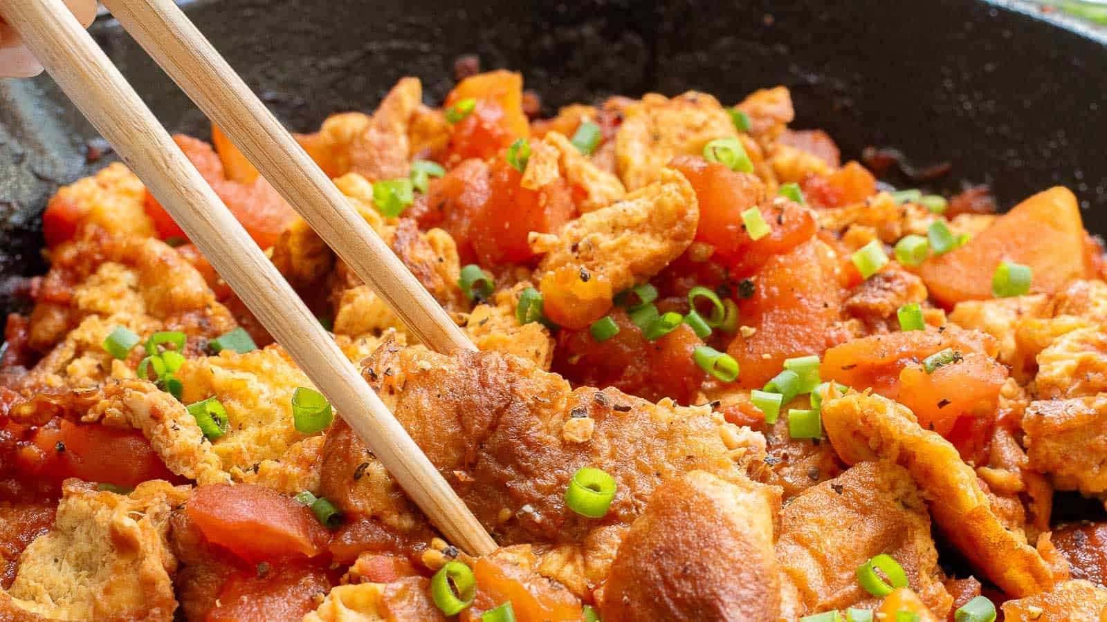 Chopsticks pick up a portion of scrambled eggs stir-fried with tomatoes and garnished with chopped green onions in a pan.