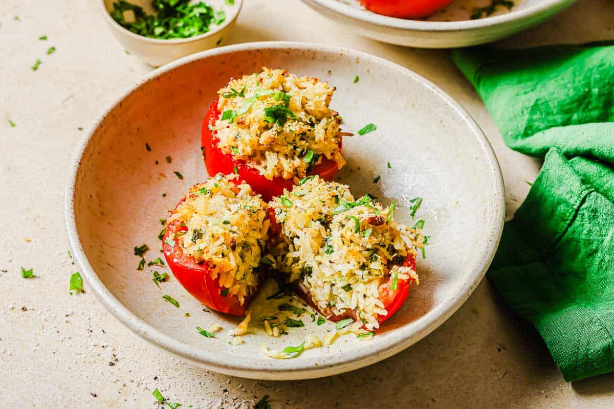 Three halved tomatoes stuffed with rice and herbs are served on a beige plate, garnished with chopped parsley. A green napkin and a bowl of herbs are nearby.