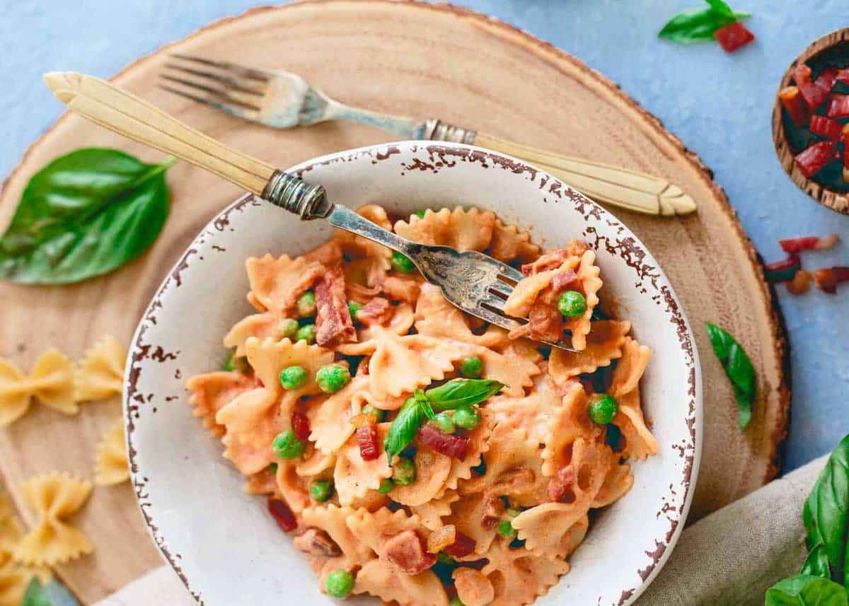 A bowl of farfalle pasta with peas, bacon pieces, and a creamy sauce, garnished with fresh basil leaves, sits on a wooden board with a fork and knife.